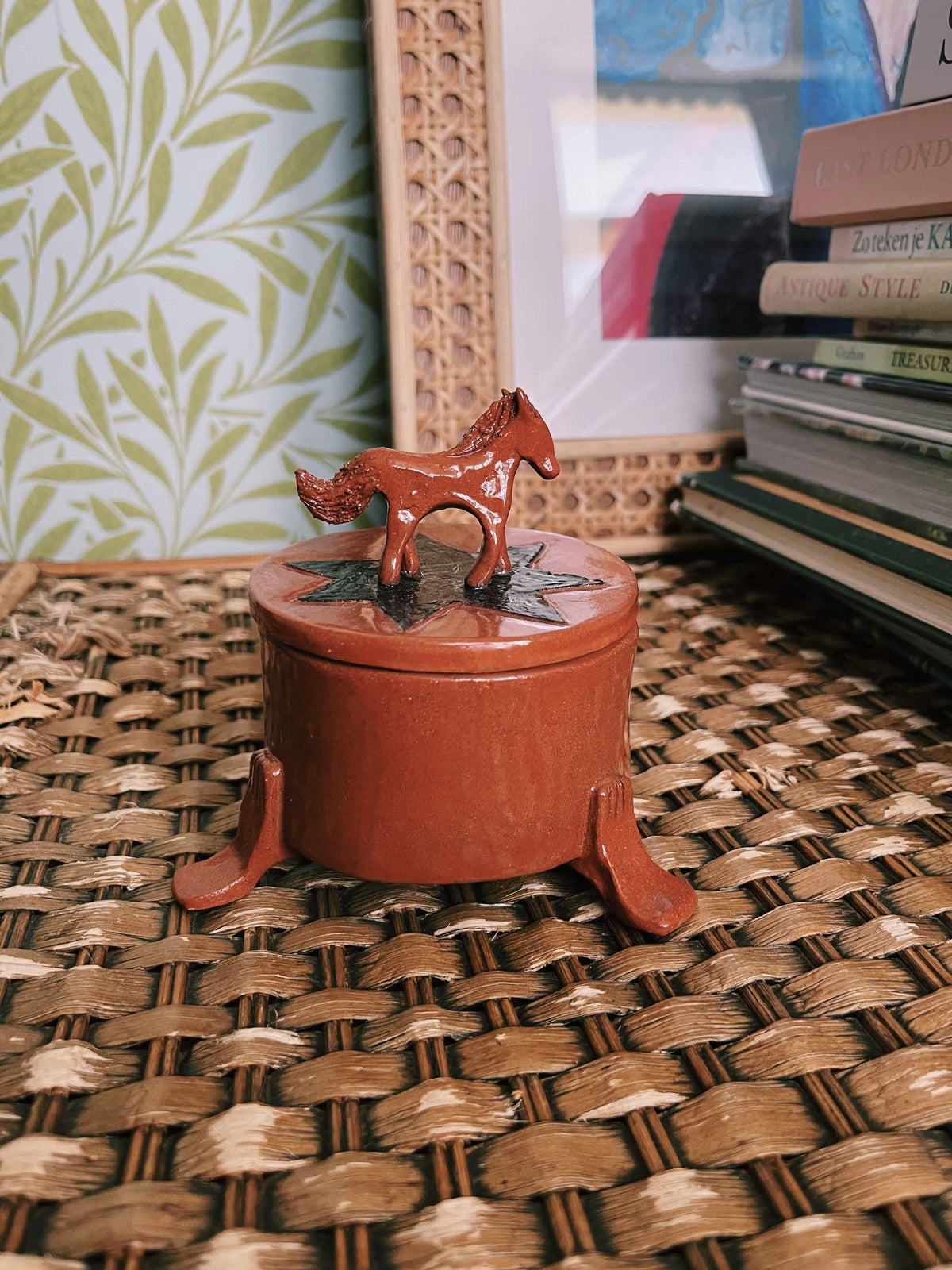 A small round terracotta-colored decorative box with a lid and a horse-shaped topper, placed on a woven wicker surface with books and wallpaper in the background.