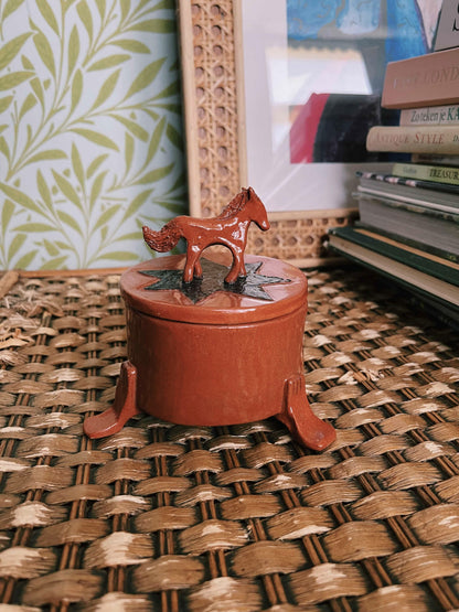 A small round terracotta-colored decorative box with a lid and a horse-shaped topper, placed on a woven wicker surface with books and wallpaper in the background.