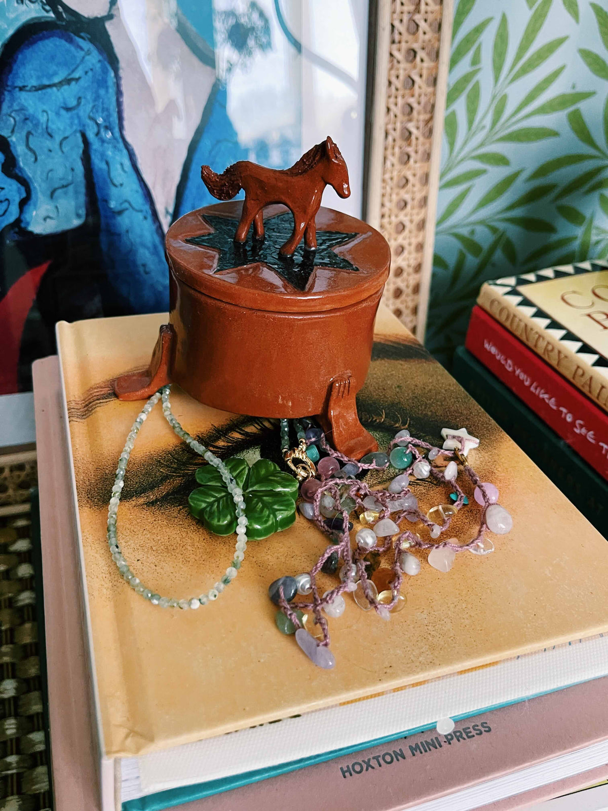 A small round terracotta-colored lidded box with a horse figurine on top, styled on top of books with beads and jewelry nearby.
