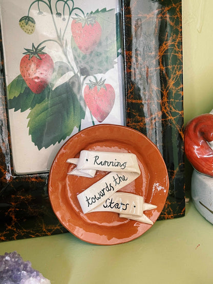 A small round terracotta-colored ceramic plate with a glossy finish, featuring a cream-colored ribbon decoration that reads 'Running towards the Stars'.