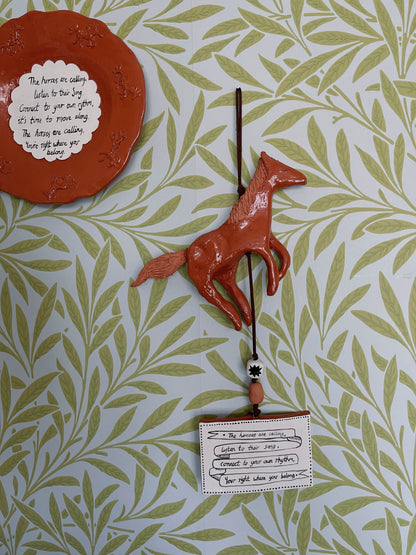 A terracotta-colored ceramic horse wall hanging suspended from a string with a small hanging card, displayed against a green and white patterned wallpaper background.