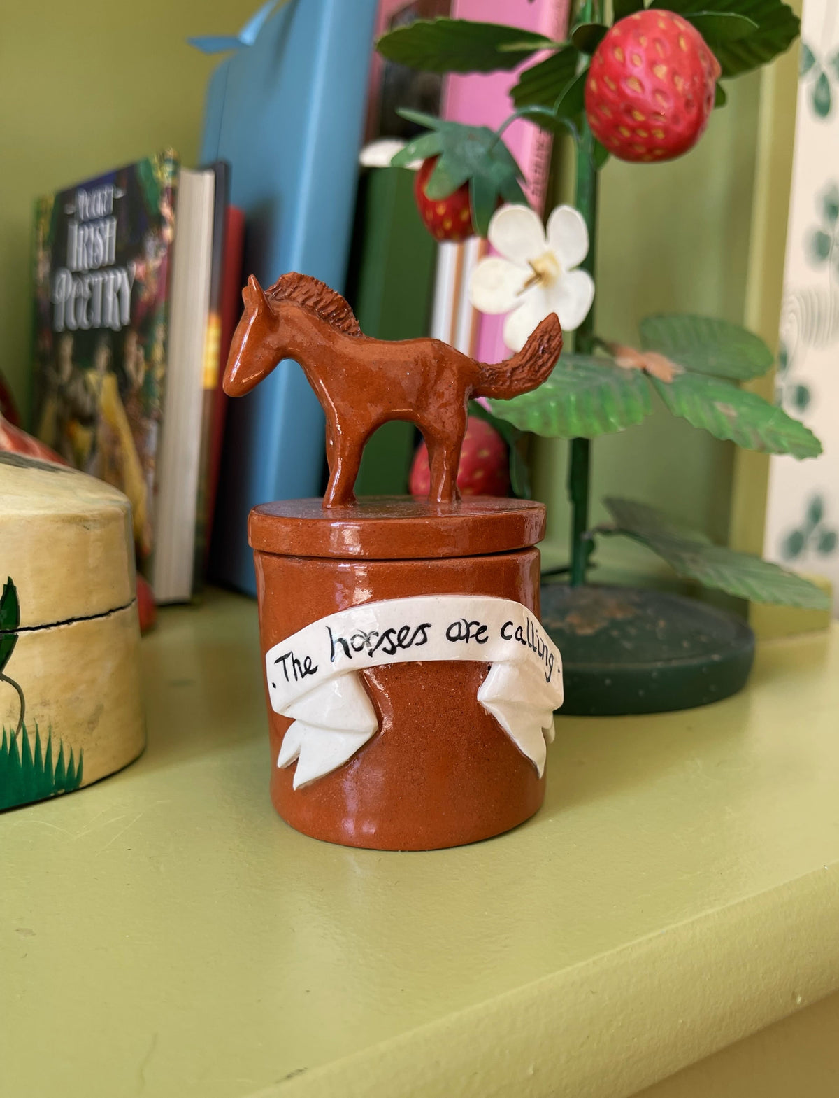 A small brown ceramic horse figurine standing on top of a matching cylindrical container, with a white ribbon decoration that reads 'The horses are eating'. The item is displayed on a shelf in a home setting with books 