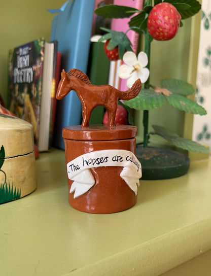 A small brown ceramic horse figurine standing on top of a matching cylindrical container, with a white ribbon decoration that reads 'The horses are eating'. The item is displayed on a shelf in a home setting with books 