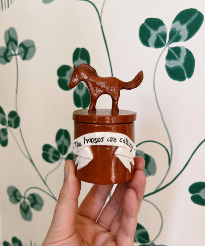 A hand holds a small brown ceramic joss stick holder featuring a horse figurine on top and a white ribbon decoration with the text 'The horses are calling'. 