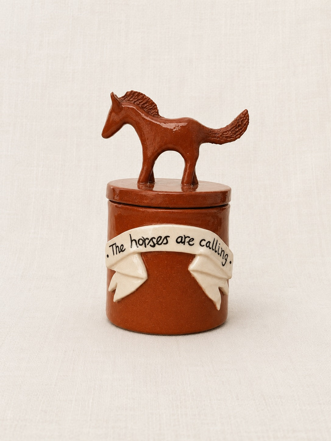 A small brown ceramic lidded container with a horse-shaped lid topper and a cream ribbon decoration around the front that reads 'The horses are calling.