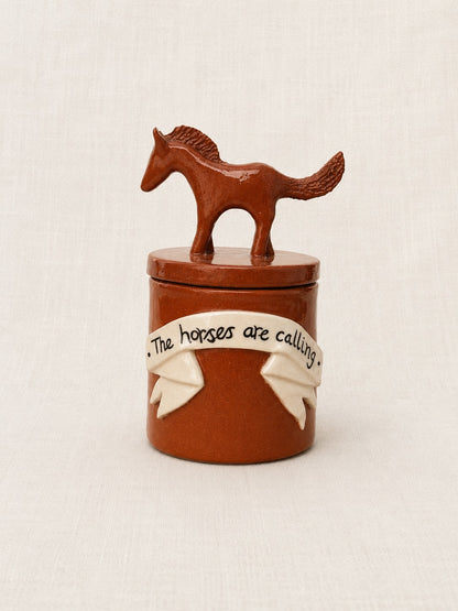 A small brown ceramic lidded container with a horse-shaped lid topper and a cream ribbon decoration around the front that reads 'The horses are calling.