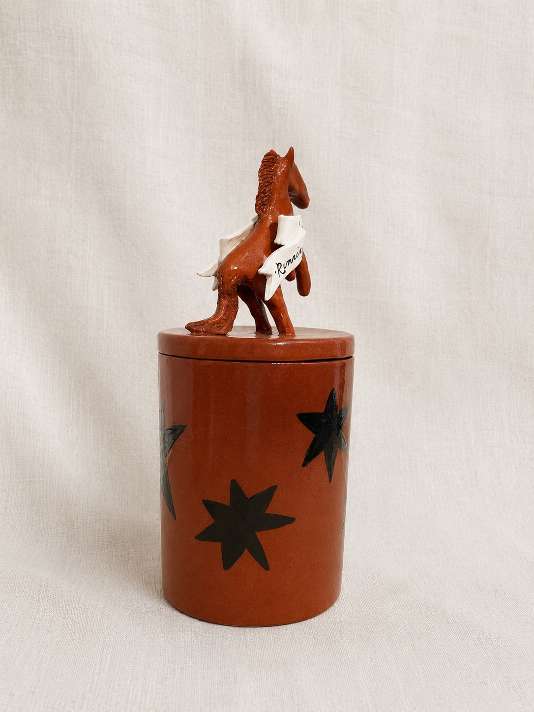 A brown cylindrical lidded container with black star cutouts, topped with a small brown horse figurine wearing a white sash, set against a light fabric backdrop.